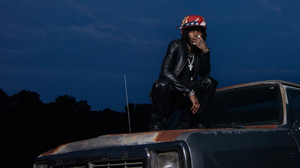 Rapper J.I.D posing, seated on the top of a car.
